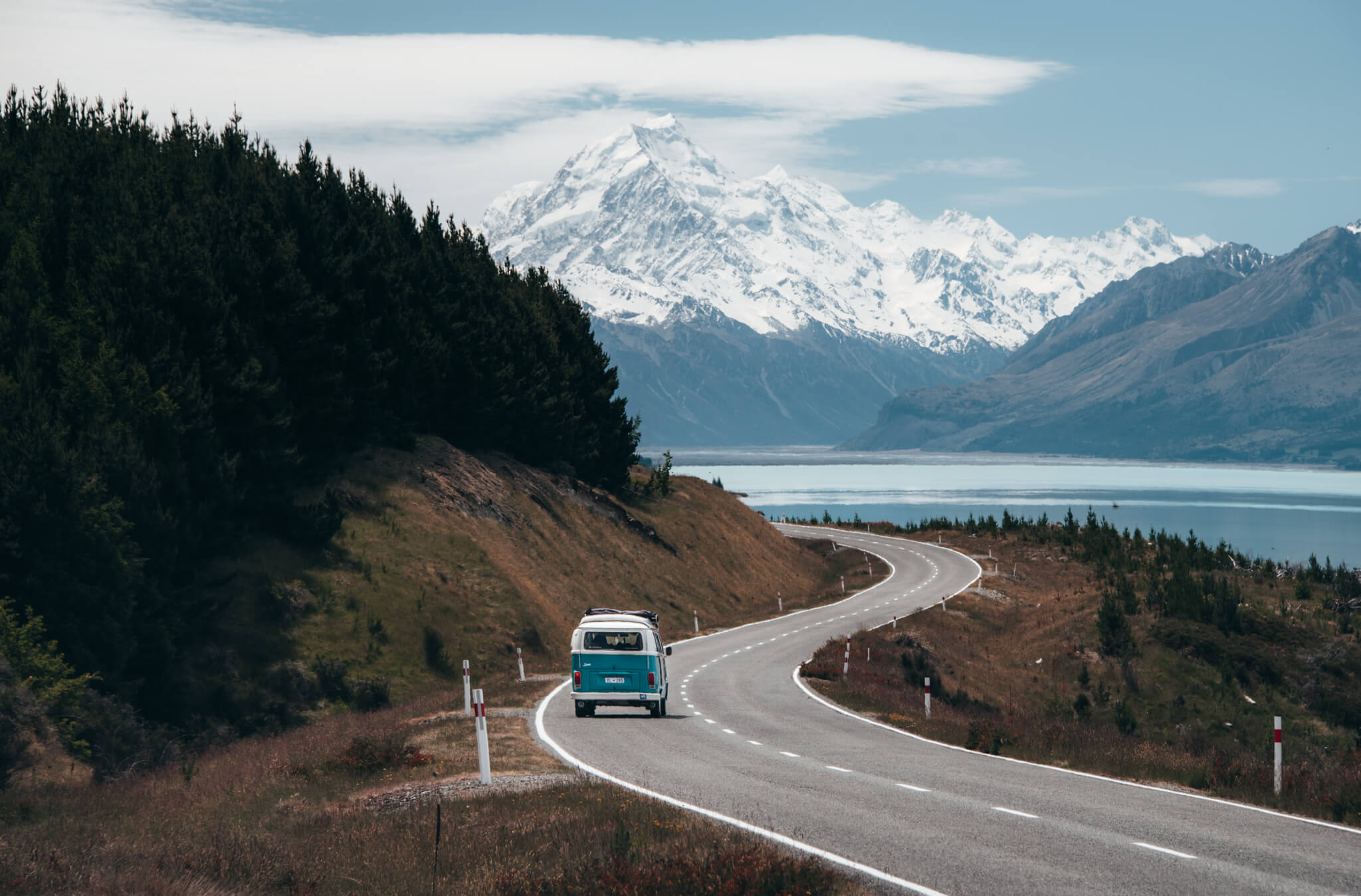 Blue van driving on a winding road by forest and lake with snow-capped mountains in the background.